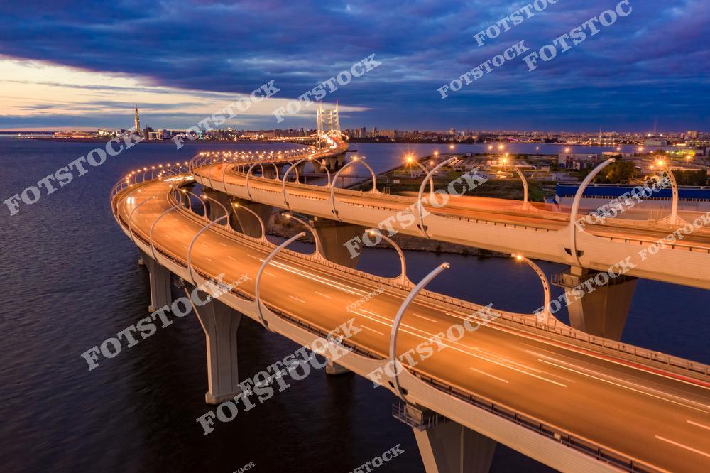 На фотографии изображен Вантовый мост, который является частью современной транспортной инфраструктуры Санкт-Петербурга. Яркие огни освещают дороги и мости, создавая динамичную картину вечернего города. Вдалеке виднеются небоскрёбы и другие здания, подчеркивающие развитие города. Снимок с высоты передает как инженерные достижения, так и атмосферу города на фоне реки.